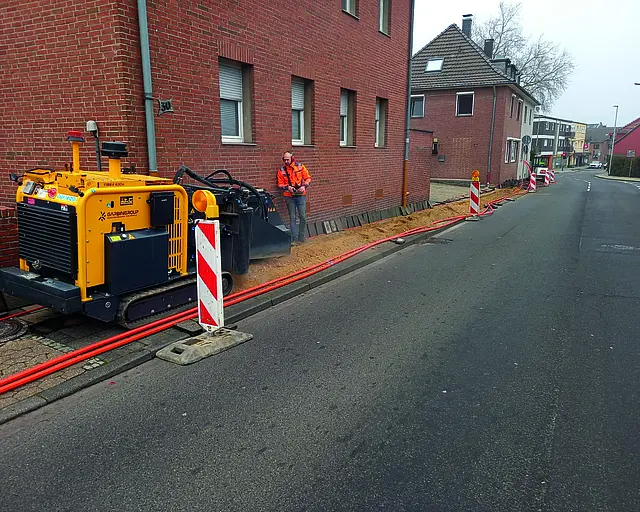 Neben einer Hauswand wird ein Graben gezogen, ein Baugerät ist zu sehen.