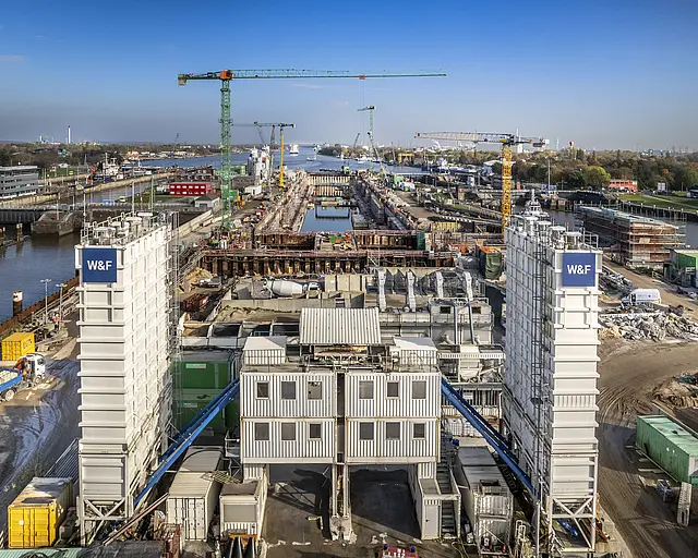 Blick auf den Bau der 5. Schleusenkammer in Brunsbüttel, man sieht Kräne und eine Wasserbaustelle, sowie die blauen Unternehmenslogos im Vordergrund
