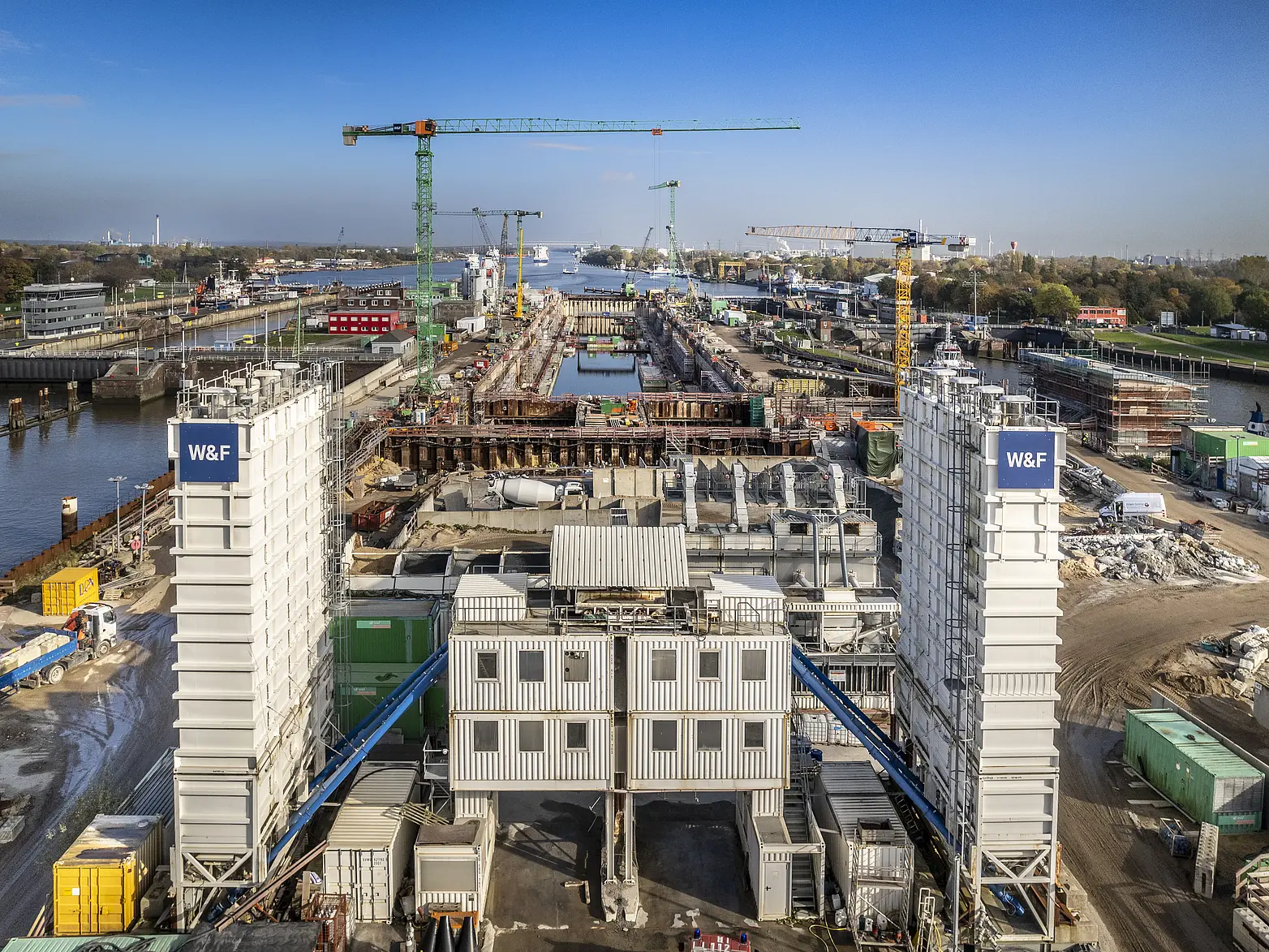 Blick auf den Bau der 5. Schleusenkammer in Brunsbüttel, man sieht Kräne und eine Wasserbaustelle, sowie die blauen Unternehmenslogos im Vordergrund